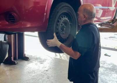 Auto mechanic in a service bay removing a front wheel from a red car lifted on a hoist at C&J Automotive.