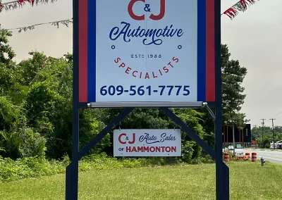Roadside sign for C&J Automotive & Tire of Hammonton featuring their name, establishment year 1988, phone number 609-561-7775, and a smaller sign below set against grass and trees along the roadway.