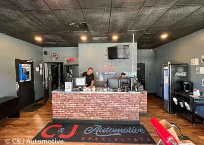 Front desk area at C&J Automotive of Newtown Square with staff working behind a brick counter, logo wall and TV above and a refreshment station.