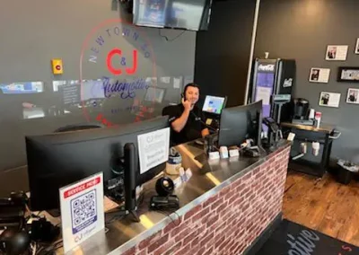 Service advisor at C&J Automotive & Tire of Newtown Square speaking on the phone behind the front counter with computer monitors and the shop logo on the wall.