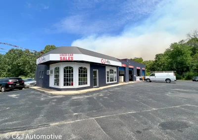 Exterior view of C&J Automotive’s office and adjacent multi-bay auto repair shop, with large front windows, service bay signs, parked vehicles, and a wide paved lot.