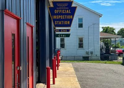 Entrance of C&J Automotive with red service bay doors, a red safety bollard row, and a hanging Pennsylvania Official Inspection Station sign.