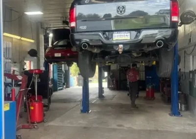Interior of C&J Automotive Specialists' service bay with a black pickup truck raised on a lift for undercarriage repair, a technician working nearby, and additional vehicles and tools visible throughout the shop.