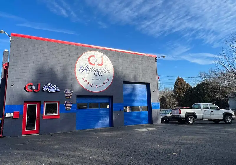 C&J Automotive & Tire in Parkesburg, a gray auto repair building with a large circular C&J logo, bright blue service bay doors, official inspection station signs, and vehicles parked alongside the shop.