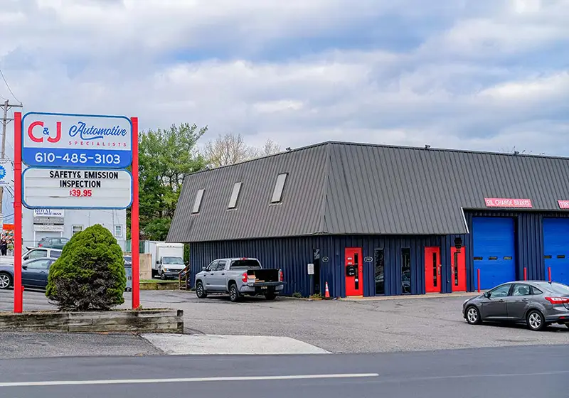 C&J Automotive in Upper Chichester with a roadside sign advertising safety and emissions inspections, a navy blue repair shop with red doors and blue service bays, and customer vehicles parked outside.