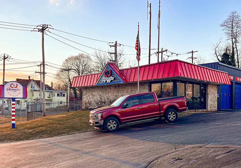 Exterior of C&J Automotive Specialists in Berwyn featuring a stone-front building with a red metal roof, blue service bay doors, a roadside business sign, and a red pickup truck parked out front.