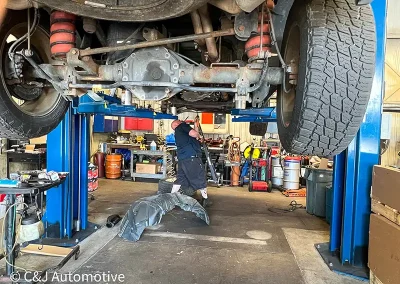 Technician at C&J Automotive inspecting the undercarriage of a truck raised on a lift, showing the exposed axle, suspension, and tires, with a wheel liner removed and shop tools in the background.